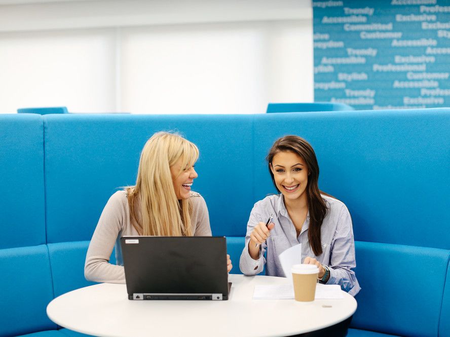 Two women sitting a blue booth laughing beside a laptop