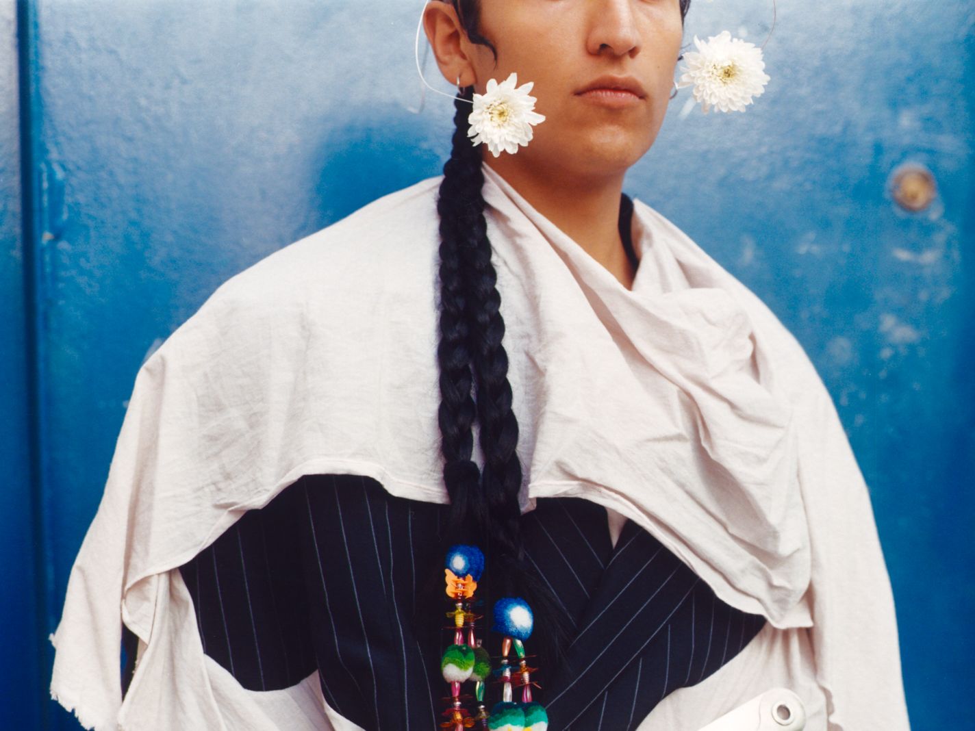 Photograph of LGBTQ+ person wearing flowers in their ears and and a robe held together with a giant clip against a blue wall. 