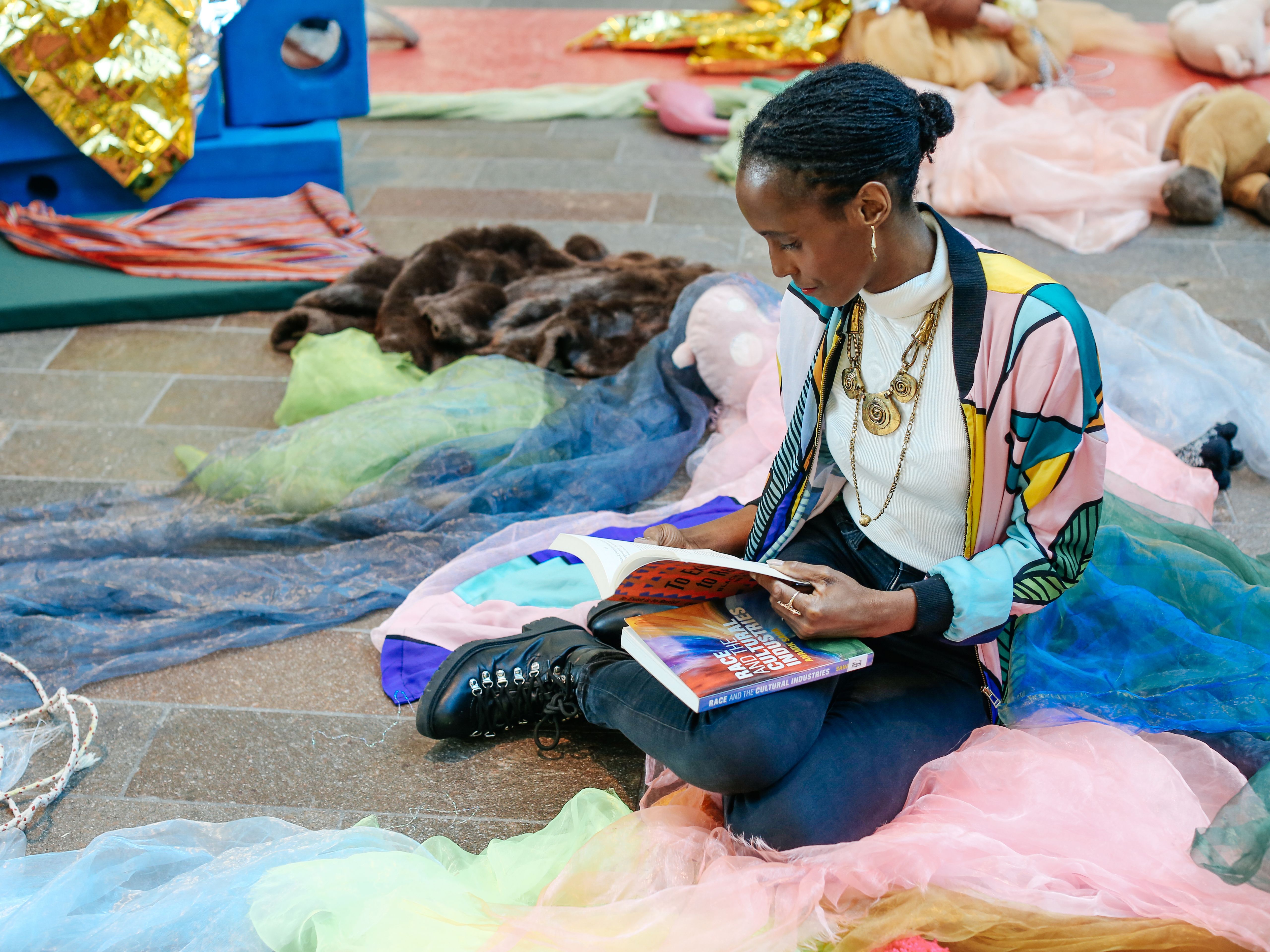Black woman sitting on the floor, reading a book surrounded by colourful fabric