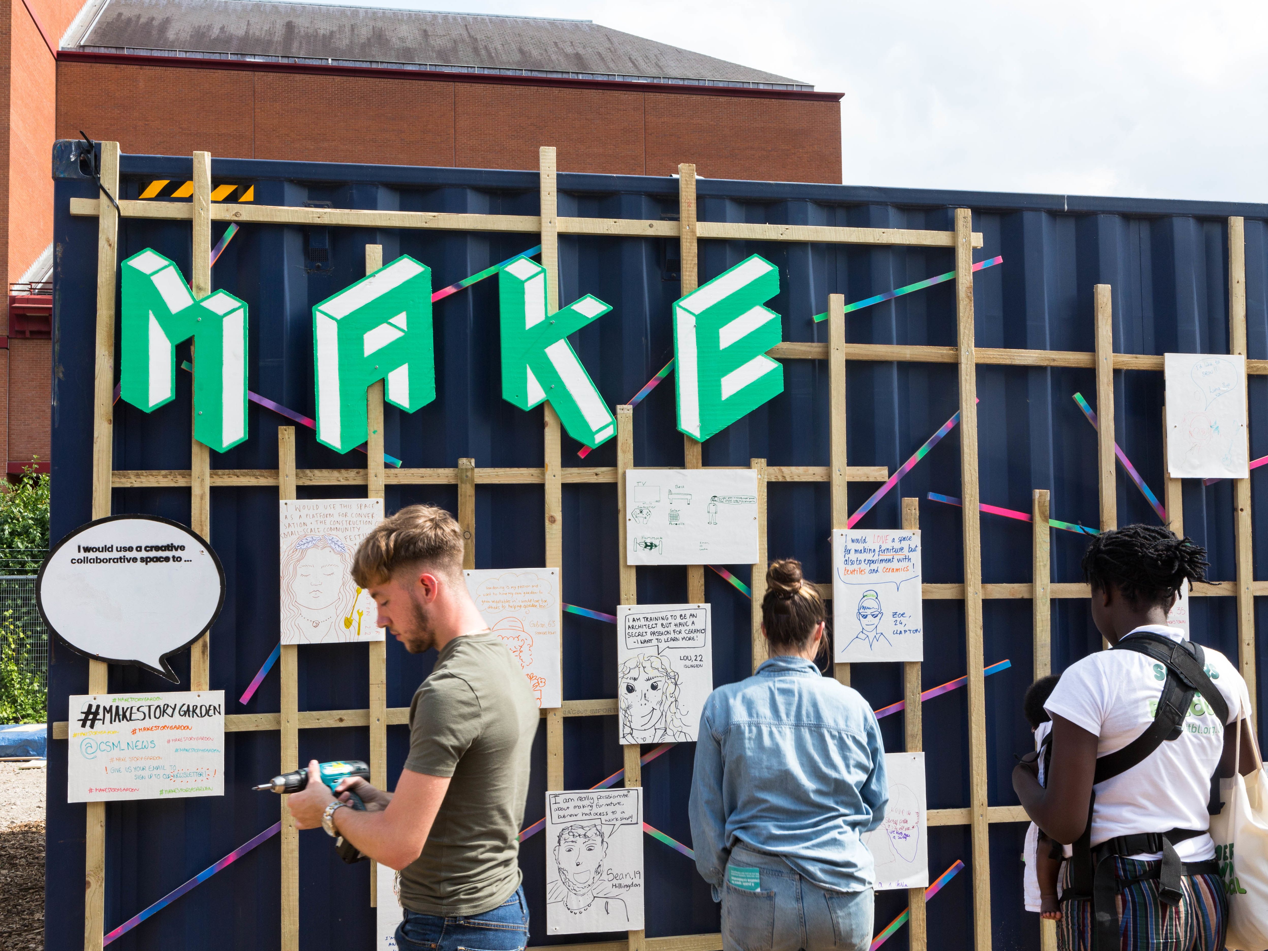A group of people are stood in front of some wooden fencing - affixed to the wood are large green letters which read 
