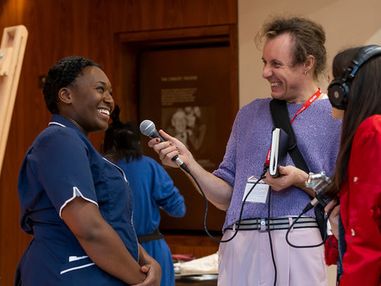A group of three people are talking. On the left and indivdual is speaking into a micraphone, they are wearing a nurses unniform. Two others stand around them