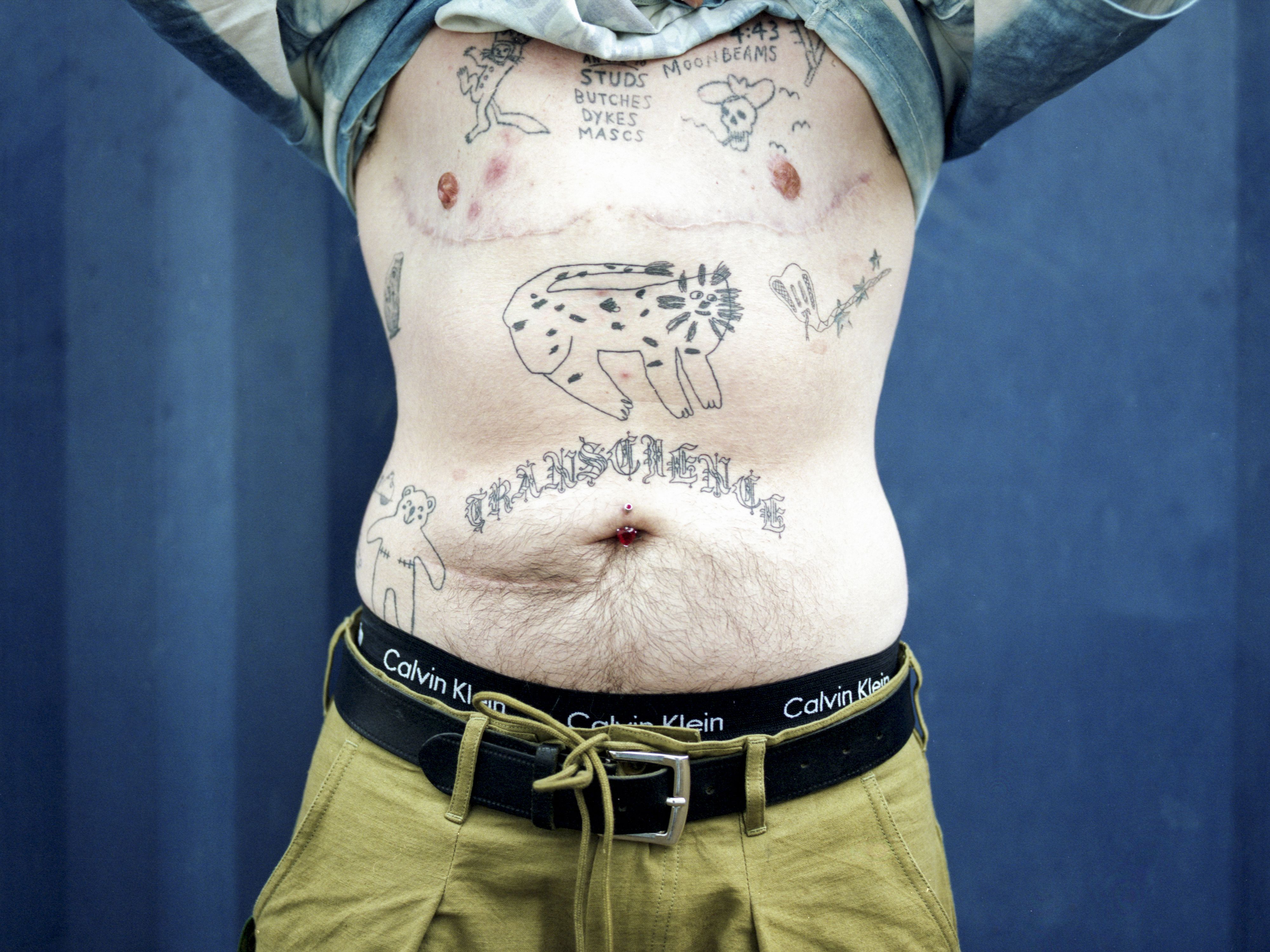 A person raises their shirt to reveal a chest covered in tattoos. The tattoos are of a doodle-like, cartoonish style.