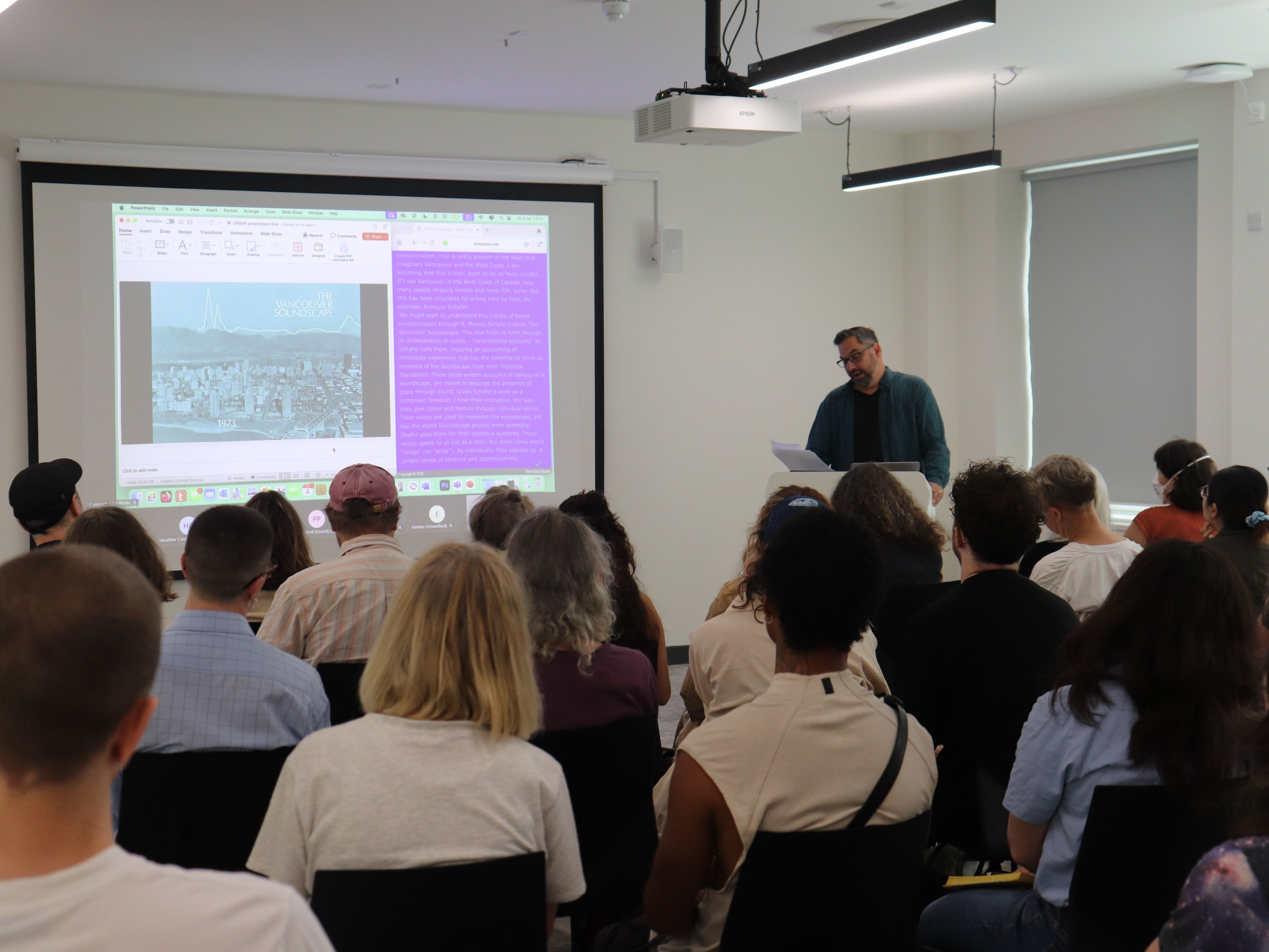 Dylan Robinson by projected slides at lectern reading from paper. Backs of audience heads.