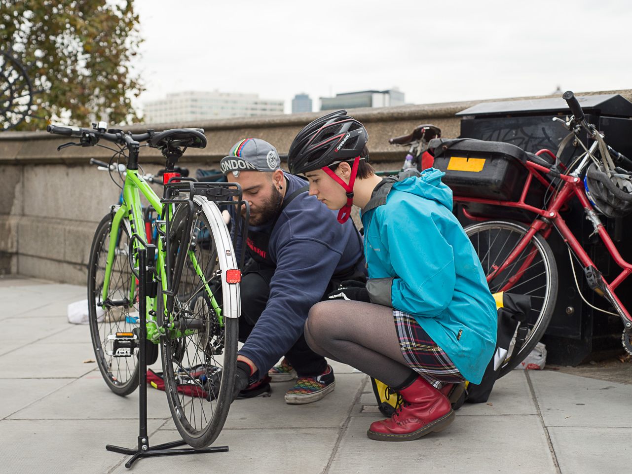 A mechanic helps a young student repair their bike on the pavement of Lambeth Bridge in London. The student is wearing a helmet and listening intently.