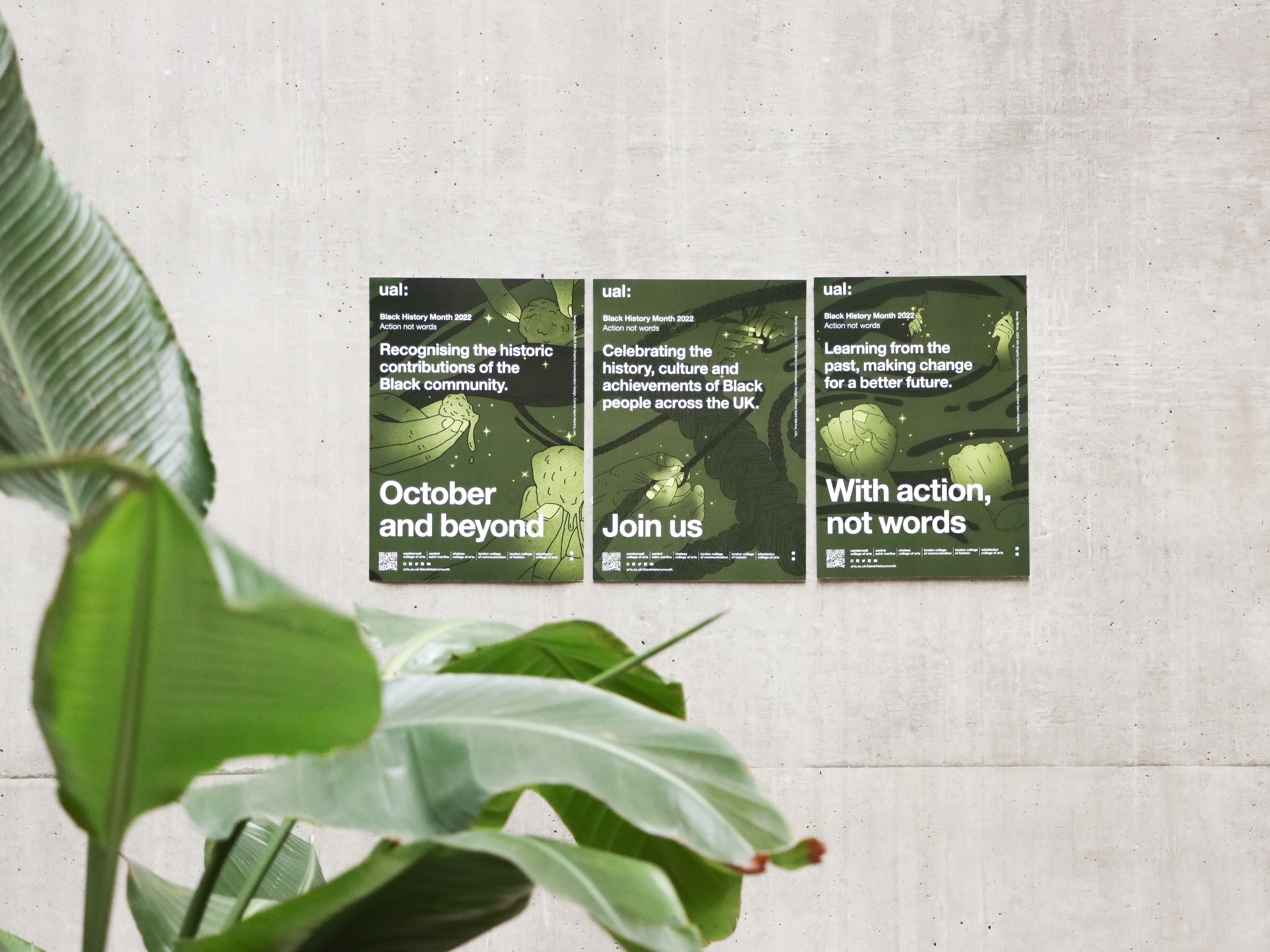 Three posters displayed on a concrete wall promoting UAL Black History Month 2022. The posters highlight themes such as recognizing the contributions of the Black community, celebrating culture and achievements, and advocating for action over words. Large green leaves frame the scene in the foreground.