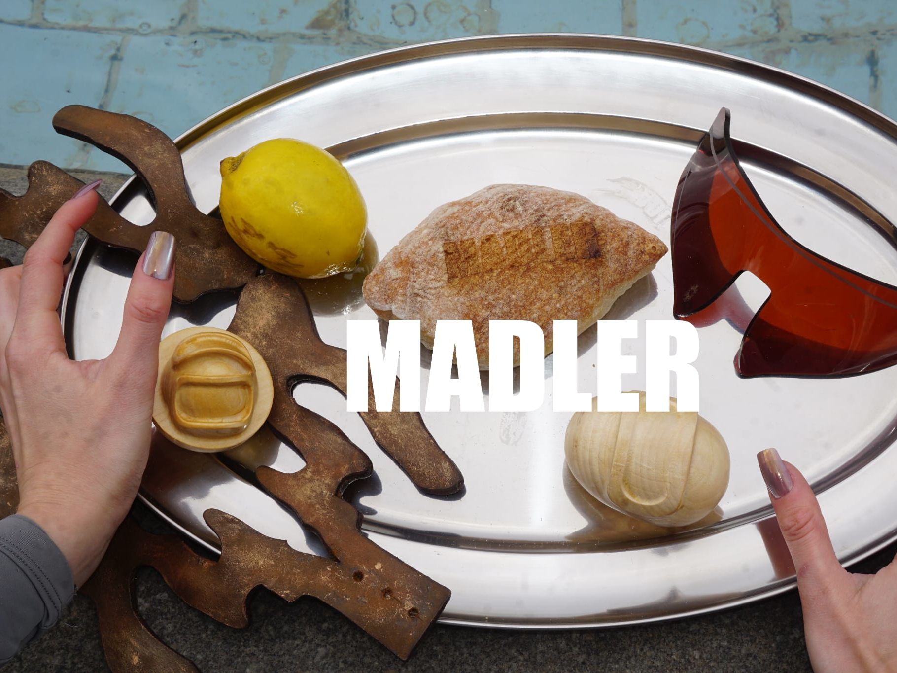 Two hands holding a silver platter with a lemon, piece of bread, leather cutouts, wooden balls, and a pair of sunglasses.