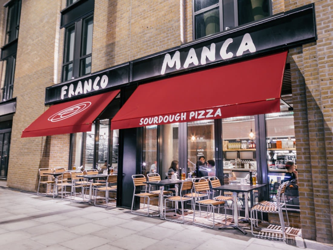 A red banner which says Franco Manca  hands above the door of a brick building. People are sat outside eating and drinking.