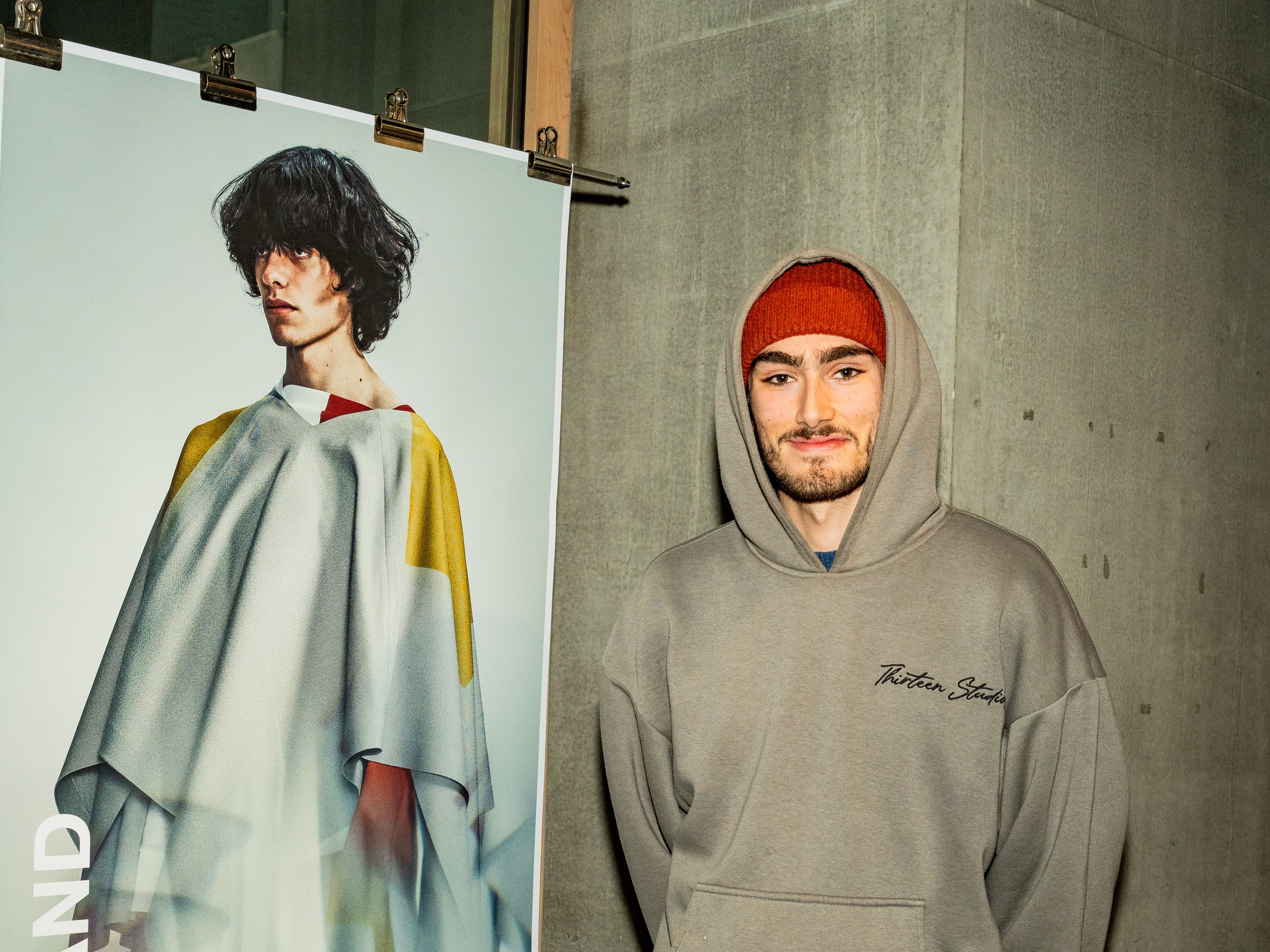 Person smiling at camera, next to roll down poster of model dressed in grey garment.