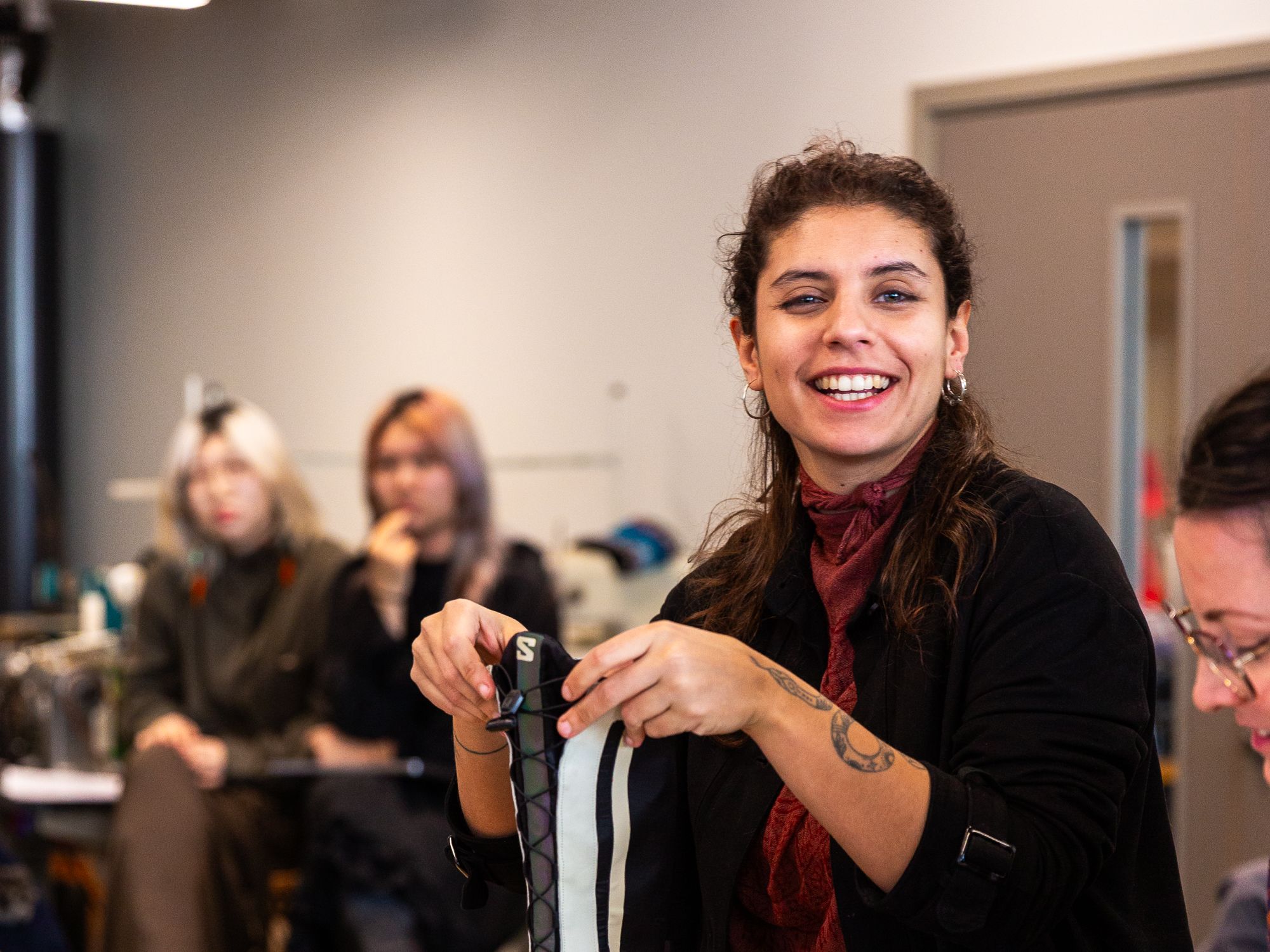 A woman holding a boot and smiling towards the camera with two people in the distant background and another person writing with a pen.