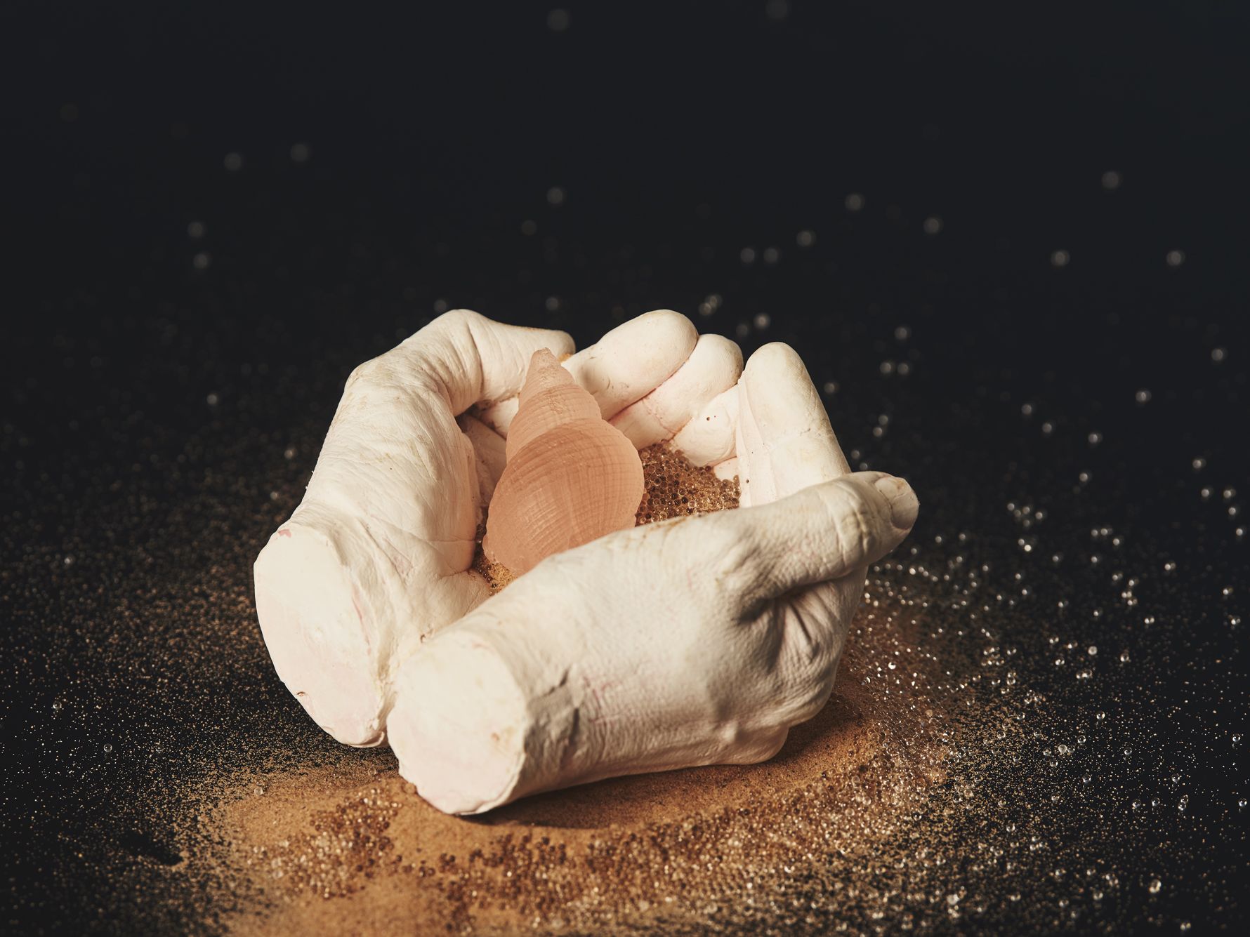 Plaster cast of a pair of hands holding a pink opaque shell and sand. 