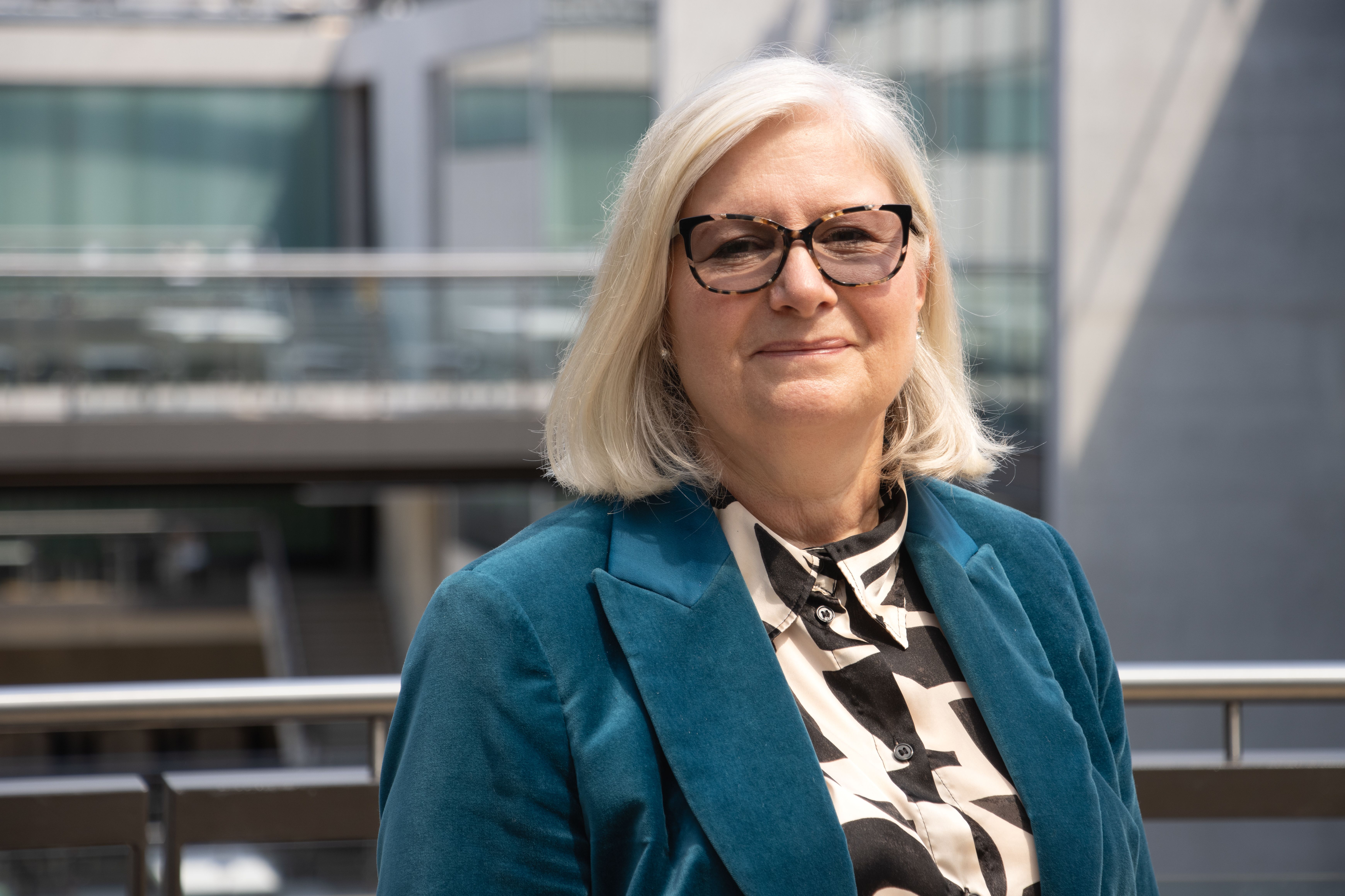 A landscape colour portrait of Sally McGill, shot against an aluminium railing with a glass-fronted space in the background.