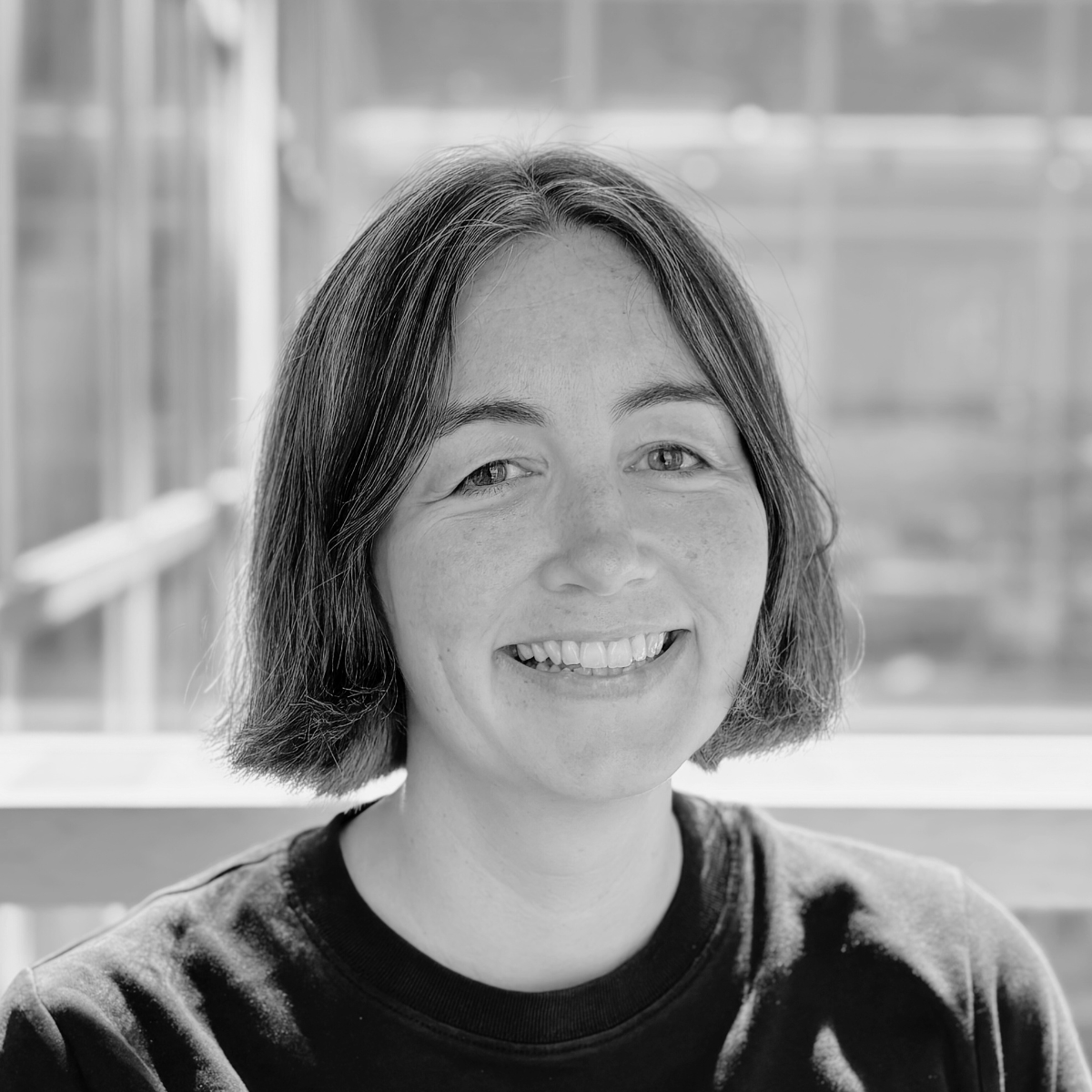 Sarah Lynch, Operations Assistant, is standing in front of a white wall wearing a black top, she has clear rimmed glasses and short black hair, the image is black and white. 