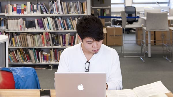 Photo of a man siting at a table in the library using his laptop and reading a book