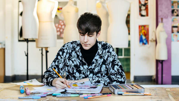Male student sitting at desk drawing in sketchbooks, with mannequins behind him