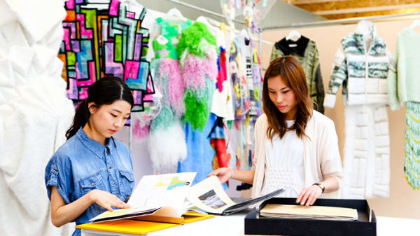 Photo of two female students looking through books on a table with clothes hanging in the background