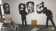 Photo of two people standing in front of a wall with paintings of faces on it