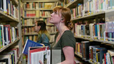 Photo of a woman holding books, looking at a bookcase