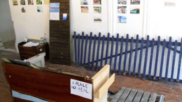Photo of a room with a blue fence leaning against the wall as well as am upturned desk with a sign on saying