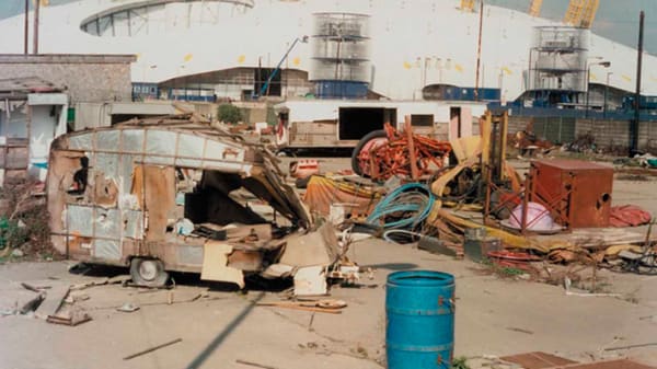 A still from Professor William Raban's film MM. Wasteland space outside of the Millennium Done. Objects and a discarded caravan an oil cylinder in colourful colours, in a sunlit scene.