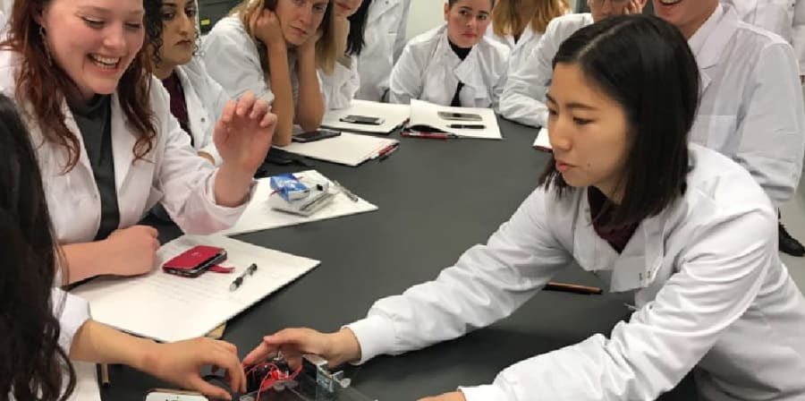 students gather round a table in a lab in white coats looking at a circuit board