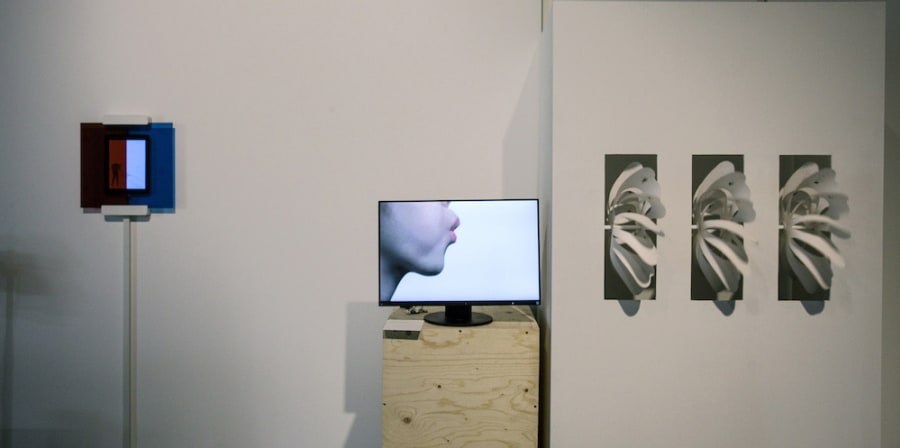 Still image of a video installation of a mouth blowing three paper turbines on the wall beside the screen