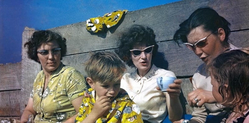 A family, wearing sunglasses, sit on a beach and drink tea.