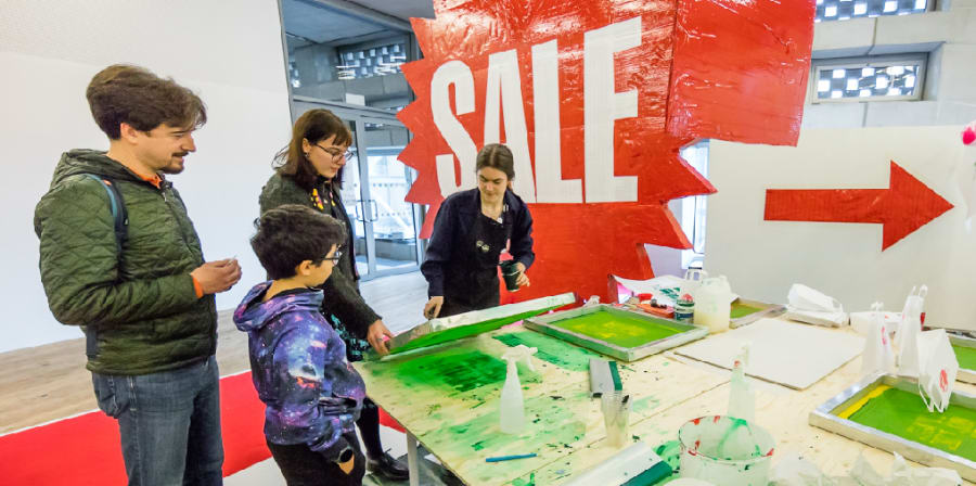 a woman screenprints money at tate exchange whilst three visitors look on, there is a banner in the background that says SALE