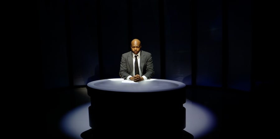 Black man in a suit sitting at a news desk in a television studio.