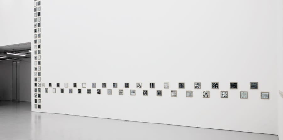 A broad white wall with an intersecting row and column of small square framed monochromatic artworks to form one work
