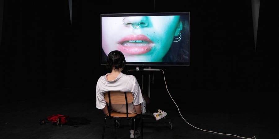 Girl watching a video installation with a close up of a girls mouth, pierced nose and ear on the screen. Work by Eva Mateos Rodriguez - BA Contemporary Theatre and Performance