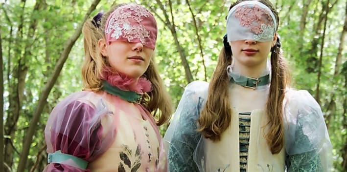 two masked models face the camera while in a forest looking away from the camera wearing garments inspired by the pre-Raphaelite art movement