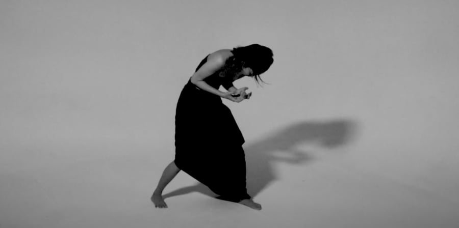 A greyscale shot of a barefoot person in a long black dress leaning forward, their hair obscures their face