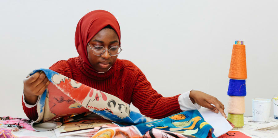 Black female student sitting at workbench looking at various brightly coloured fabrics