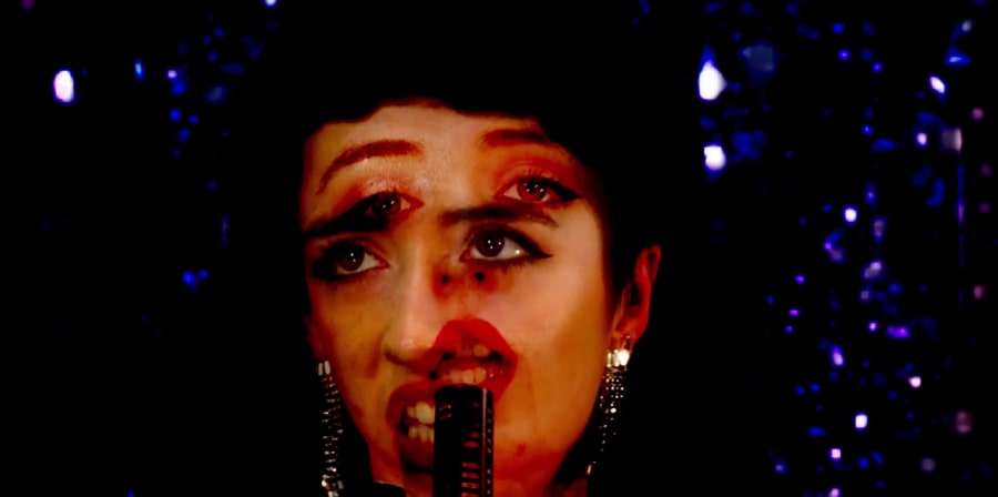 Still from video featuring someone singing into a mic in front of a sequin backdrop with their face overlaid multiple times by Katie Grenville.