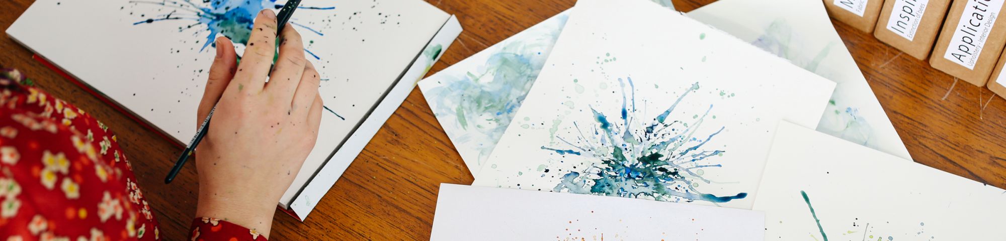 Hand with a paintbrush in and pieces of paper with blue and orange paintings on