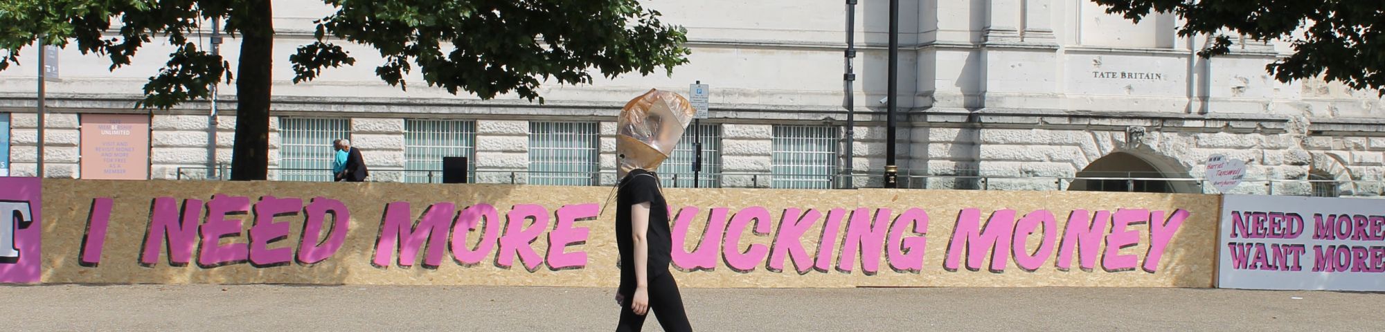 figure wearing a piece of adrienne bennie's design, behind them the fence features hand-written words: 'i need more money'