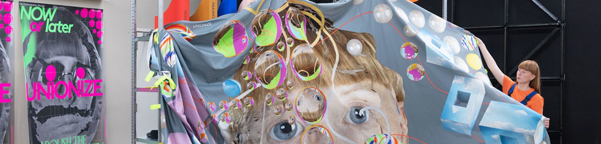 An artist stands beside a bedsheet-sized artwork depicting a face surrounded by coloured bubbles.