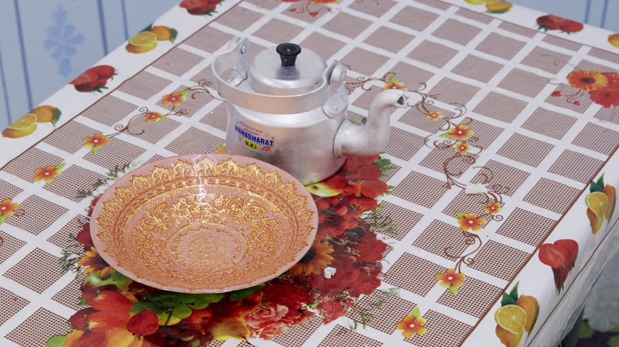 Table with decorative tablecloth with grid pattern and fruit. On the table is a metal teapot and decorative bowl. and 