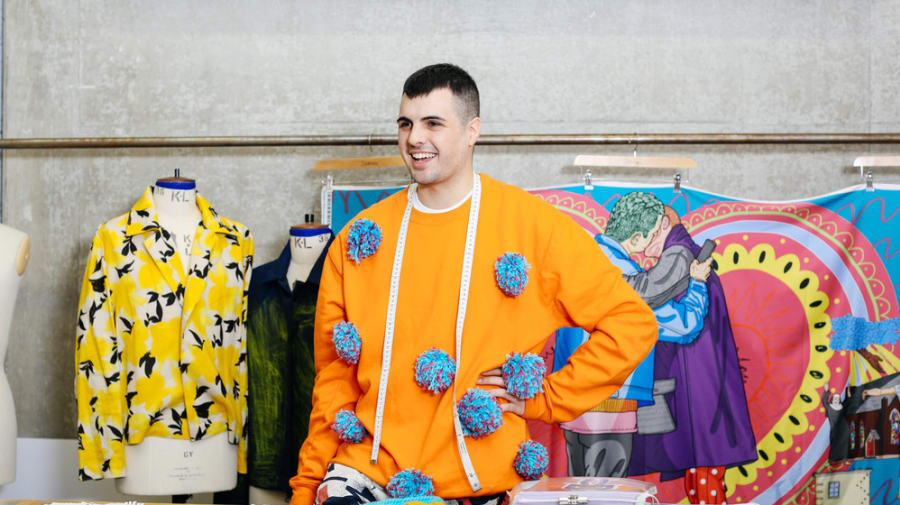 Student standing next to mannequin and behind large table with fabric samples and fashion sketches displayed.