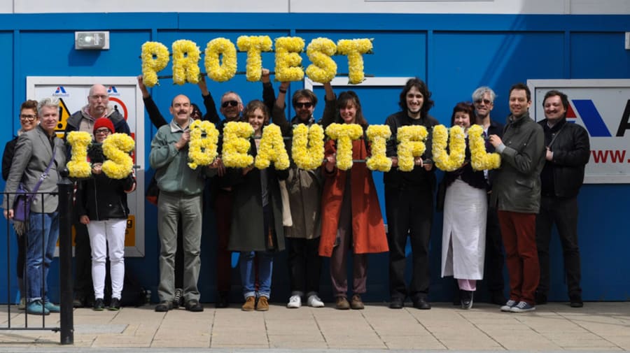Protest is beautiful banner being held up by small crowd of people.