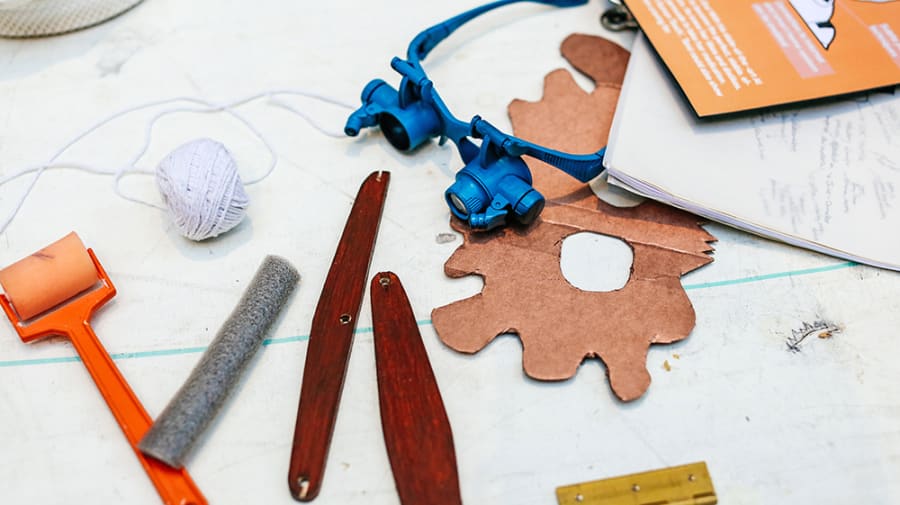 Workspace setup from Nina Cutler’s project, featuring an assortment of crafting tool - paint roller, sandpaper, wooden sticks, string, goggles, and cardboard gear - on a white surface with faint blue lines.