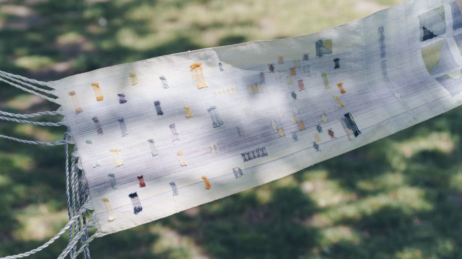 Detail of a woven textile by Anna Hashimoto, featuring a light base with multicolored patchwork and fringed edges, photographed outdoors against a grassy backdrop.