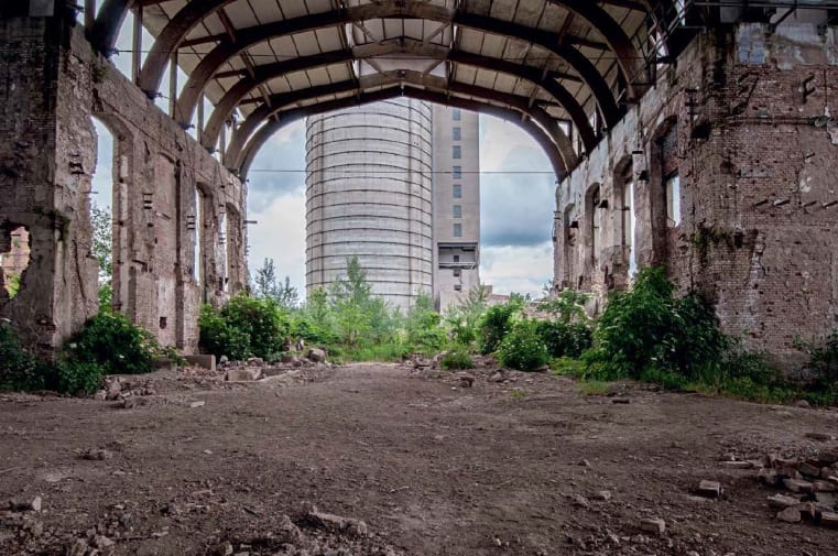 A derelict industrial building with an internal archway overgrown with grass and shrubs and a newer brutalist style building beyond the arch