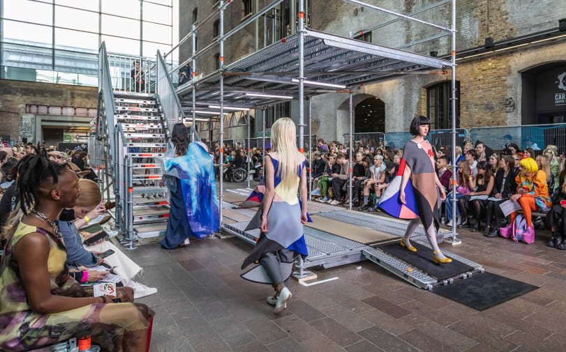 Two floor catwalk made in scaffold with three models wearing colourful garments