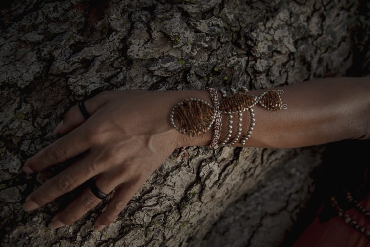 A hand adorned with multiple beaded bracelets and rings rests against a textured tree bark