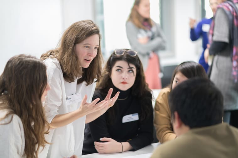 A woman in a white shirt is talking and gesticulating to a group of seated students.