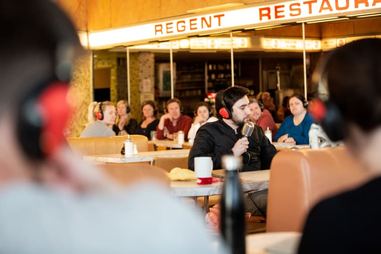 A man holds an old-fashioned microphone in his hand, sitting at a table in a full diner style restaurant.