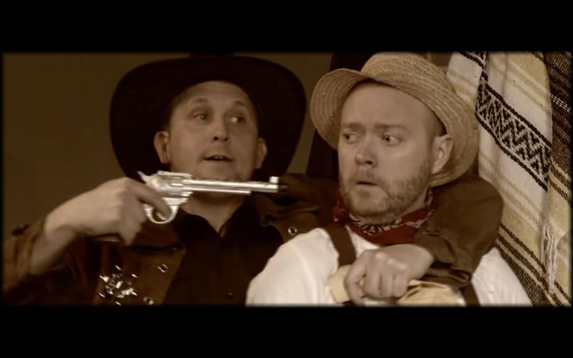 Man in cowboy-style hat holds a gun to the head of a man.