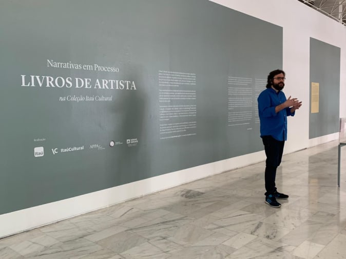 A casually dressed professor shot against a large internal grey wall with Portuguese writing 