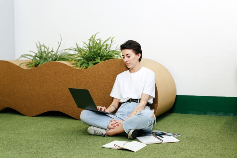 A person sits on a laptop with notebooks scattered around them. They are on astroturf with a cork planter behind them